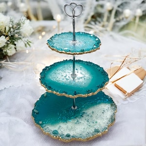 Pode incluir: Um suporte de servir de três níveis com pratos de resina estilo ágata azul-turquesa e branca. As bordas são douradas e o suporte central é prateado. A alça superior tem um design de coração decorativo. Ideal para sobremesas.