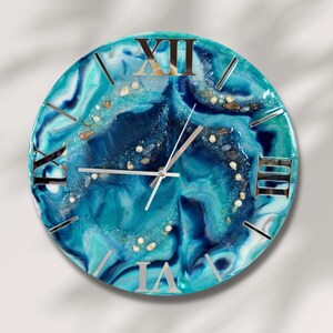 Könnte beinhalten: Runde Wanduhr mit blauem und türkisfarbenem Harzdesign, silbernen römischen Ziffern und Zeigern. Das Zifferblatt hat eingebettete dekorative Elemente und ein glänzendes Finish, was eine einzigartige Uhr ergibt.