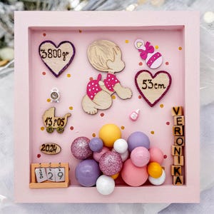 Può includere: Una cornice rosa per bambini con ritagli di legno ed elementi decorativi. Include una figura di bambino, cuori con misure (3800g, 53cm), un passeggino, un orologio e il nome "Veronika".