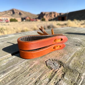 May include: Close-up of a handcrafted leather strap in shades of brown and tan. The strap features two layers of leather with decorative cutouts and a small, attached leather accent. The item is set against a rustic, outdoor backdrop.