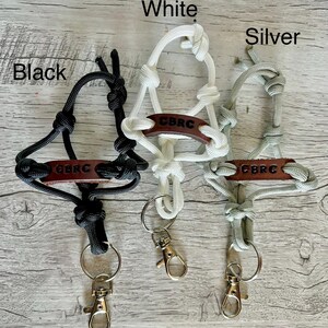May include: Three lanyards made of black, white, and silver paracord, each featuring a brown leather tag embossed with the text CBRC. Each lanyard includes a metal split ring and a lobster claw clasp, with the colors labeled as Black, White, and Silver.