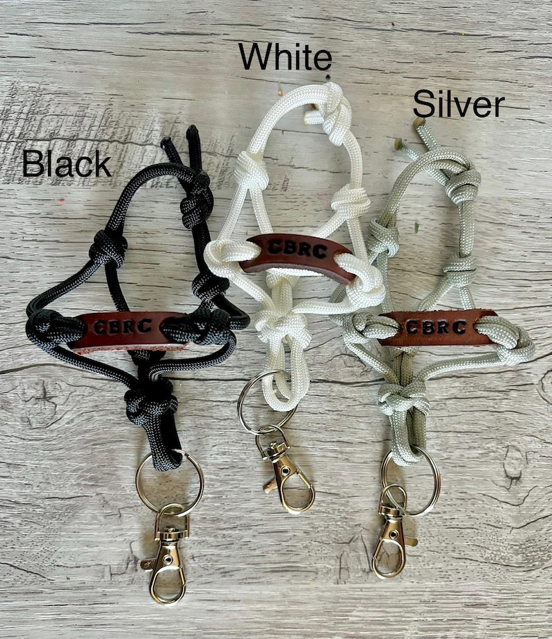 May include: Three lanyards made of black, white, and silver paracord, each featuring a brown leather tag embossed with the text CBRC. Each lanyard includes a metal split ring and a lobster claw clasp, with the colors labeled as Black, White, and Silver.