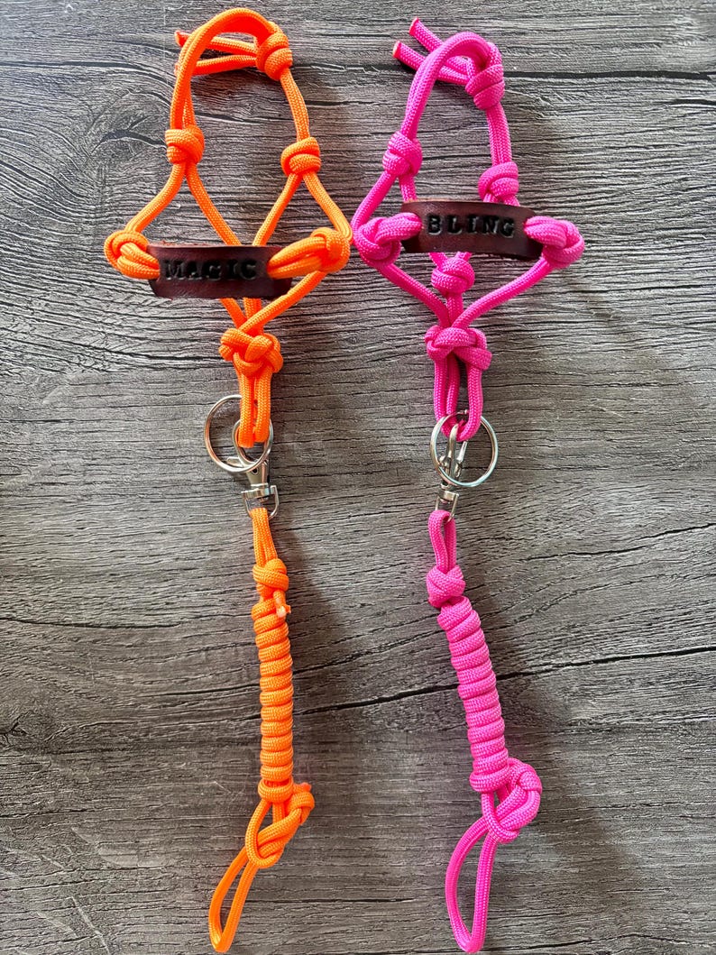 May include: Two paracord lanyards featuring neon orange and neon pink knotted cord designs. Each lanyard includes a dark brown leather tag with the words MAGIC and BLING embossed in black, a silver metal split ring, and a swivel lobster clasp.