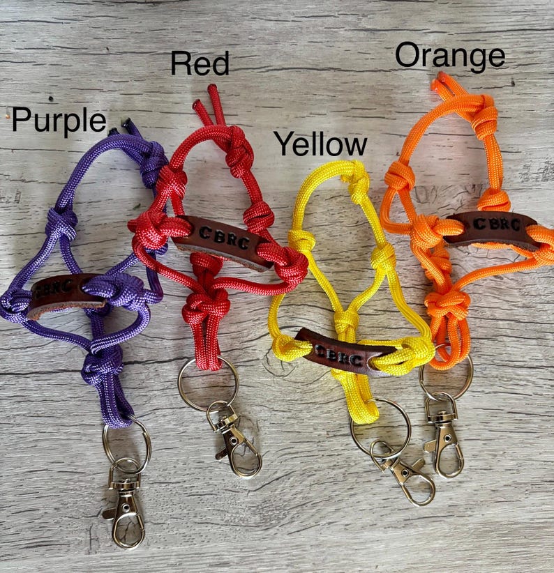 May include: Four paracord lanyards in purple, red, yellow, and orange, each featuring a brown leather tag embossed with the text CBRC. Each lanyard includes a knotted cord design, a metal split ring, and a silver-toned metal lobster clasp.