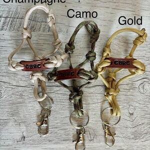 May include: Three paracord lanyards in champagne, camo, and gold colors. Each features a brown leather tag embossed with the text CBRC, knotted cord loops, a metal split ring, and a silver-toned swivel lobster clasp for secure attachment.