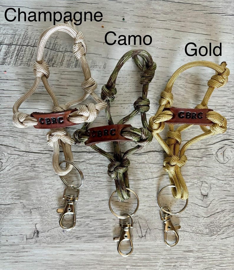 May include: Three paracord lanyards in champagne, camo, and gold colors. Each features a brown leather tag embossed with the text CBRC, knotted cord loops, a metal split ring, and a silver-toned swivel lobster clasp for secure attachment.
