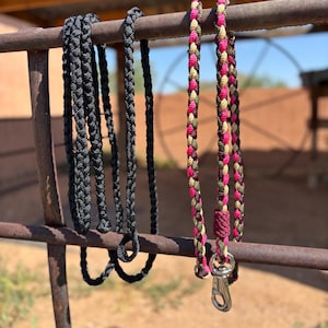 Puede incluir: Dos correas de cuerda trenzada colgadas de una barra de metal. Una correa es de color negro sólido con un extremo en bucle. La segunda correa presenta un patrón trenzado multicolor en tonos rosa, verde oliva y beige, con un mosquetón giratorio de metal plateado.
