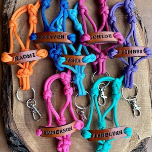May include: A collection of seven colorful paracord keychains, each featuring a brown leather tag embossed with a unique name: Naomi, Lakayen, Asia, Chloe, Timber, Emmerson, and Jadelynn. Each keychain includes a metal split ring and a lobster clasp.