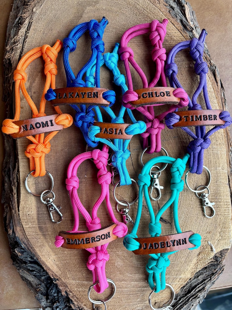 May include: A collection of seven colorful paracord keychains, each featuring a brown leather tag embossed with a unique name: Naomi, Lakayen, Asia, Chloe, Timber, Emmerson, and Jadelynn. Each keychain includes a metal split ring and a lobster clasp.