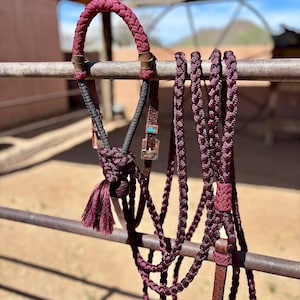 May include: A maroon braided rope horse halter with a matching long lead rope. The halter features a decorative tassel, black rope accents, and metal buckles with small turquoise-colored inlays. The braided cord is a deep burgundy color throughout.