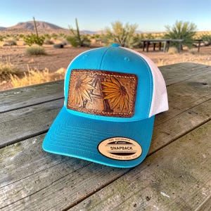 Puede incluir: Gorra snapback turquesa con parte trasera de malla blanca. La gorra presenta un parche de cuero marrón con dos girasoles tallados. El parche es rectangular y está cosido en la parte delantera. La gorra tiene una visera curva y el texto "THE AUTHENTIC SNAPBACK YUPOONG SINCE 1974".