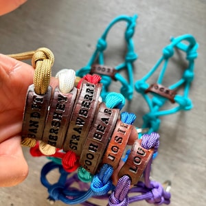 May include: A collection of colorful paracord rope halters featuring brown leather nameplates. The leather tags are stamped with the names BANDIT, HERSHEY, STRAWBERRY, POOH BEAR, JOSIE, and LOU. The ropes are in shades of gold, white, red, blue, and purple.