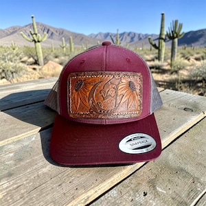 May include: A burgundy trucker hat with a leather patch featuring a floral design. The hat has a mesh back and a curved brim. The patch is brown with a sunflower and moon design. The hat has a snapback closure.