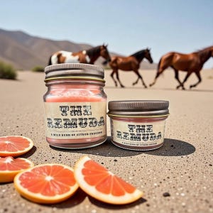 May include: Two glass jar candles with metal lids, labeled "THE REMUDA, A WILD RUSH OF CITRUS FREEDOM," sit next to grapefruit slices. The candles have a pink hue. Horses are in the background, suggesting a Western theme.