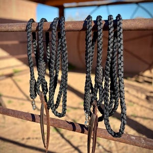 Puede incluir: Dos riendas de caballo trenzadas en negro y gris colgadas de una barra de metal oxidado. Cada rienda tiene una borla de cuero y un cierre plateado. El fondo muestra un entorno soleado al aire libre.