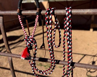 May include: A braided rope halter and lead set featuring a tri-color pattern of black, white, and red. The halter includes a solid black noseband, brown leather accents, and a decorative tassel knot at the chin, with a matching braided lead rope.