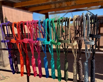 May include: A collection of colorful rope horse halters in various shades, including purple, orange, pink, turquoise, green, tan, gray, and black. The halters are hanging on a metal bar, with a rustic wooden structure in the background.