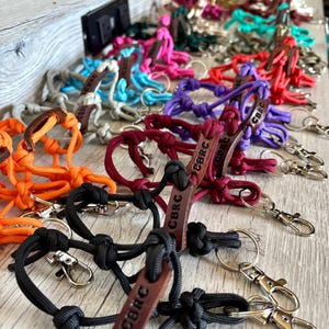 May include: A collection of multi-colored paracord rope keychains featuring intricate knots and a brown leather tag embossed with the letters CBRC. Each keychain includes a silver-toned metal swivel lobster clasp and a circular split ring attachment.