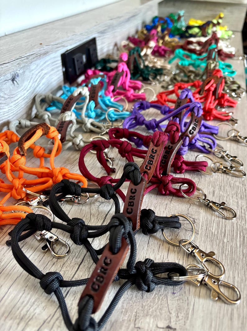 May include: A collection of multi-colored paracord rope keychains featuring intricate knots and a brown leather tag embossed with the letters CBRC. Each keychain includes a silver-toned metal swivel lobster clasp and a circular split ring attachment.