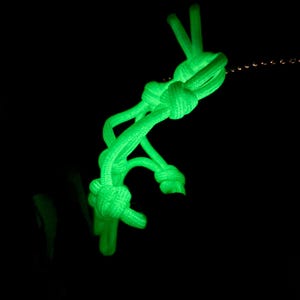 May include: A bright neon green paracord keychain featuring a series of intricate, tightly woven knots. The cord glows intensely against a solid black background, highlighting the textured surface of the braided nylon material and the knot structure.