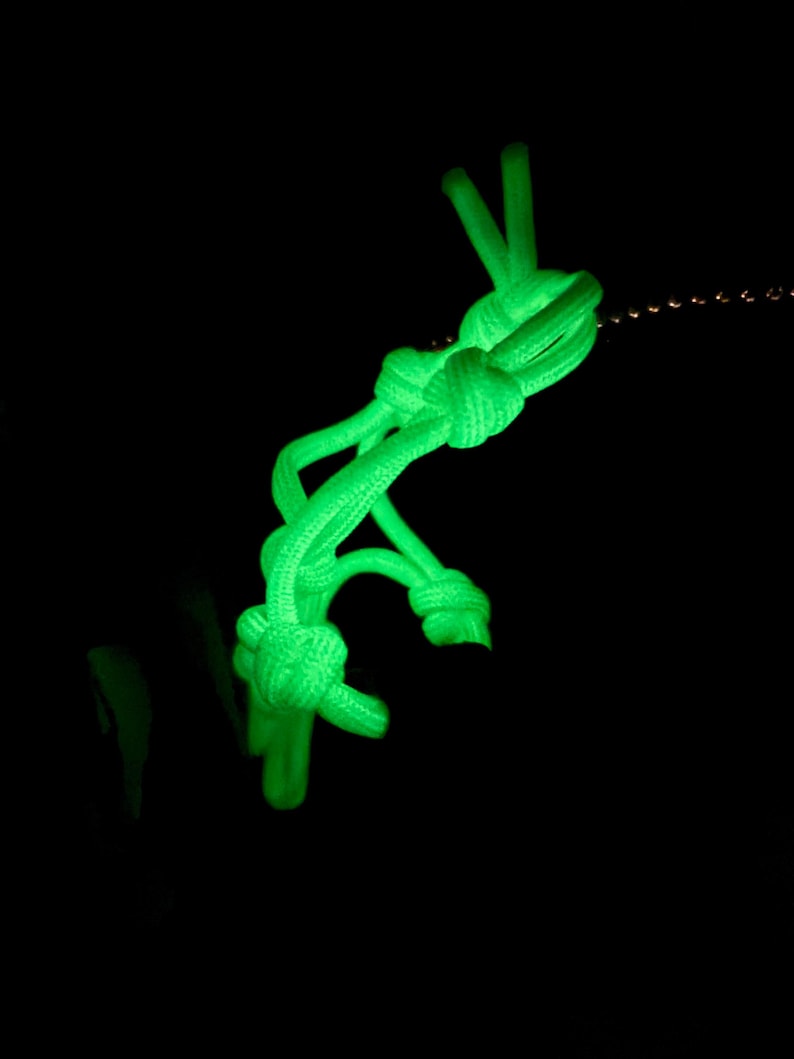 May include: A bright neon green paracord keychain featuring a series of intricate, tightly woven knots. The cord glows intensely against a solid black background, highlighting the textured surface of the braided nylon material and the knot structure.