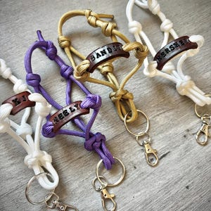 May include: Four paracord keychains in white, purple, gold, and white, each featuring a dark brown leather tag stamped with names like KJ, NEELY, BANDIT, and HERSHEY. Each keychain includes a metal split ring and a lobster claw clasp.