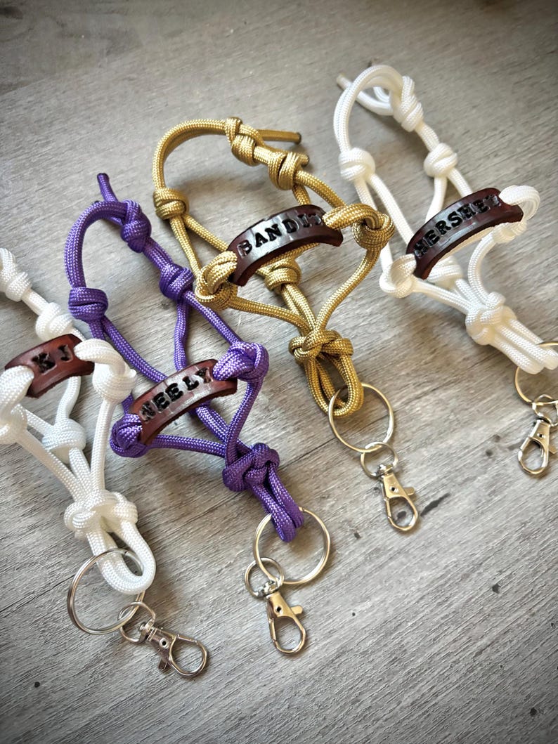May include: Four paracord keychains in white, purple, gold, and white, each featuring a dark brown leather tag stamped with names like KJ, NEELY, BANDIT, and HERSHEY. Each keychain includes a metal split ring and a lobster claw clasp.