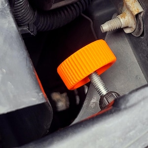 Op de afbeelding: Close-up van een oranje plastic knop met een metalen schroefdraad. De knop heeft een geribbeld oppervlak en is bevestigd aan een zwart onderdeel, waarschijnlijk onderdeel van de motor of het mechanische systeem van een voertuig.