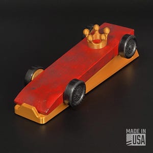 Pode incluir: Um carro de corrida de brinquedo de madeira vermelho e laranja com rodas pretas e uma coroa dourada. O carro tem um acabamento desgastado e é rotulado como "MADE IN USA".