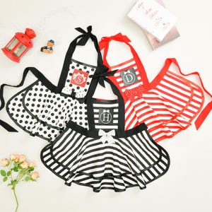 May include: A collection of aprons in various patterns, including polka dots, stripes, and solid colors. Each apron features a different initial, bow accents, and contrasting trim. The aprons are in black, white, and red.
