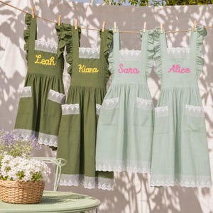May include: Four aprons hanging on a clothesline, two in olive green and two in sage green. Each apron features ruffled shoulder straps, white lace trim on the pockets and hem, and embroidered names: Leah, Kiana, Sara, and Alice in yellow or pink.