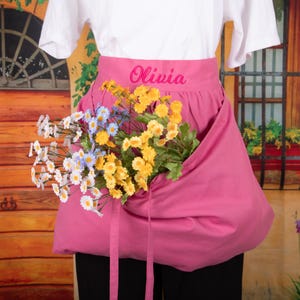 May include: A pink apron with the name "Olivia" embroidered in pink script. The apron is filled with a variety of colorful artificial flowers, including white, yellow, and blue daisies. The apron is worn over a white shirt and black pants.