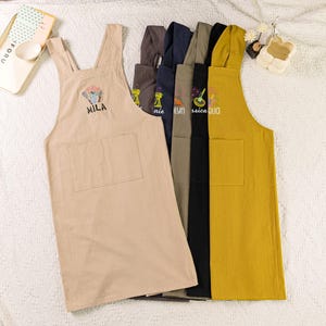 May include: Six different colored aprons with various embroidered designs. The aprons are made of a soft, linen-like fabric. The aprons are all in a neutral color palette, with the exception of one yellow apron. The aprons have adjustable straps and a large pocket on the front.