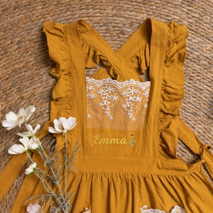 May include: Mustard yellow apron with ruffled edges and a white lace panel. The apron features the embroidered name "Emma" in a cursive font, accented with a small floral design. The apron is designed with criss-cross straps and a gathered skirt.