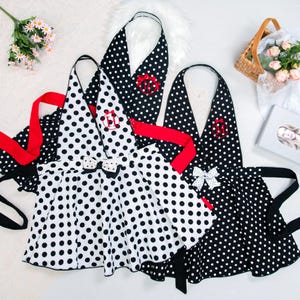 May include: Several polka dot aprons in black and white with red accents and bows. The aprons have a halter neck design and are decorated with embroidered initials. The aprons are displayed on a white surface.