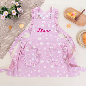 May include: A light purple apron featuring a white daisy pattern, ruffled shoulder straps, and two ruffled front pockets. The name Diana is embroidered in pink script across the chest. The apron has a cross-back design with long fabric ties.