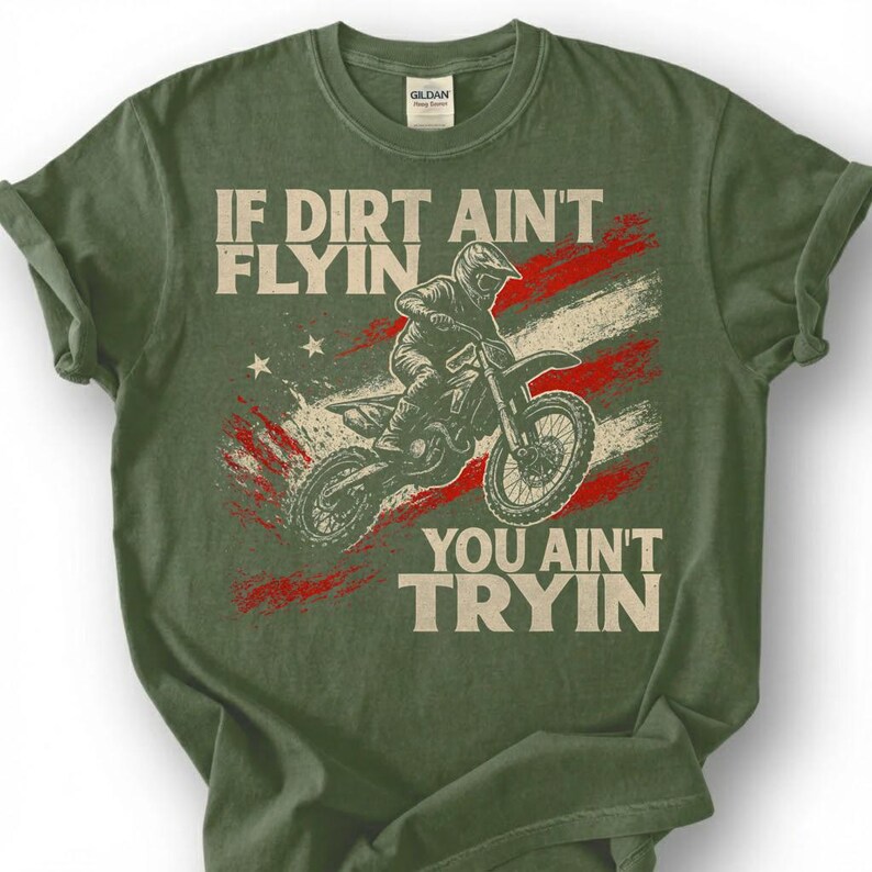 Peut inclure: T-shirt vert olive avec un motif de pilote de motocross et le texte "IF DIRT AIN'T FLYIN, YOU AIN'T TRYIN" en blanc. Le design comprend un fond rouge et blanc rappelant un drapeau.