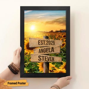 May include: Framed poster with a sunset scene over a field of sunflowers. The poster features three wooden signs with the text "EST. 2025", "ANGELA", and "STEVEN".