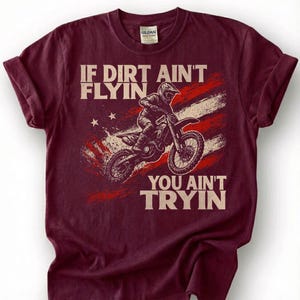 Peut inclure: T-shirt bordeaux avec un motif d'un pilote de motocross sur une moto. Le design comprend le texte "IF DIRT AIN'T FLYIN' YOU AIN'T TRYIN" avec un drapeau rouge, blanc et bleu en arri&egrave;re-plan. Le t-shirt a un col rond et des manches courtes.