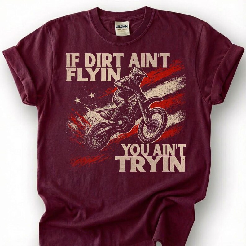 Peut inclure: T-shirt bordeaux avec un motif d'un pilote de motocross sur une moto. Le design comprend le texte "IF DIRT AIN'T FLYIN' YOU AIN'T TRYIN" avec un drapeau rouge, blanc et bleu en arri&egrave;re-plan. Le t-shirt a un col rond et des manches courtes.