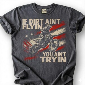 Peut inclure: T-shirt gris fonc&eacute; avec un motif de pilote de motocross sur une moto tout-terrain. Le design comprend un motif de drapeau am&eacute;ricain et le texte "IF DIRT AIN'T FLYIN YOU AIN'T TRYIN".