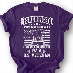 May include: A purple t-shirt with a graphic design. The design includes the text "SACRIFICED I'M NO LOSER I'M NO SUCKER I'M A U.S. VETERAN" in white, along with an American flag and a pair of combat boots.