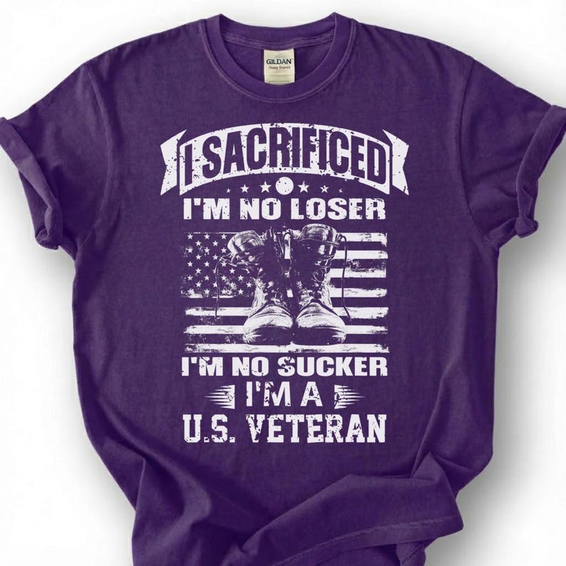 May include: A purple t-shirt with a graphic design. The design includes the text "SACRIFICED I'M NO LOSER I'M NO SUCKER I'M A U.S. VETERAN" in white, along with an American flag and a pair of combat boots.