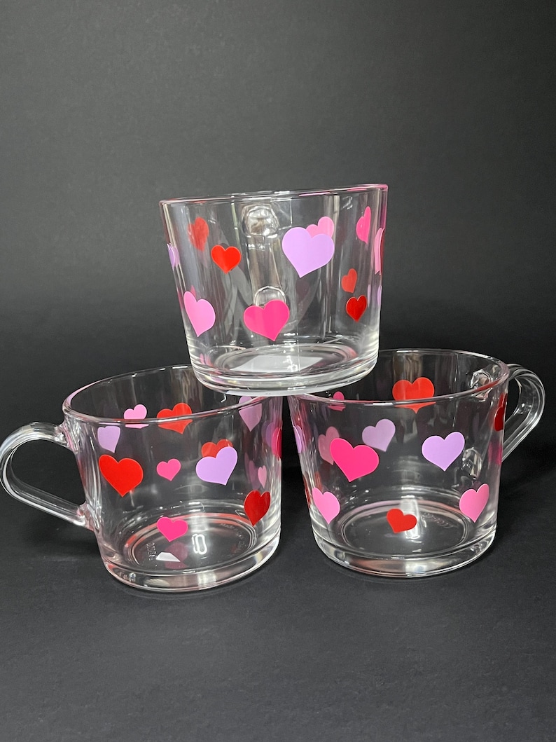 May include: Three clear glass mugs with handles, decorated with red, pink, and purple hearts. The mugs are stacked, with the top mug slightly angled. The background is a solid black.