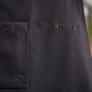 May include: Black canvas apron with a pocket and the embroidered text "goatboy" on the front.