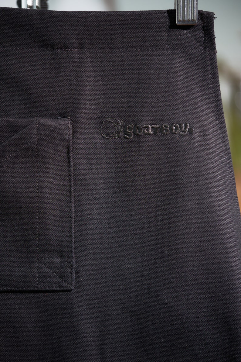 May include: Black canvas apron with a pocket and the embroidered text "goatboy" on the front.