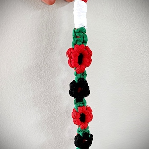 Puede incluir: Un llavero de cuerda verde, blanco y rojo con un cierre de metal negro. La cuerda está tejida en una serie de flores con centros rojos, negros y blancos.