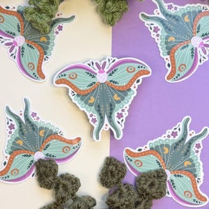 May include: Five die-cut stickers featuring a moth illustration with mint green, orange, and purple wings. The moth design includes small yellow crescent moon symbols, purple flower accents, and a pink body. The stickers are arranged on a split background.