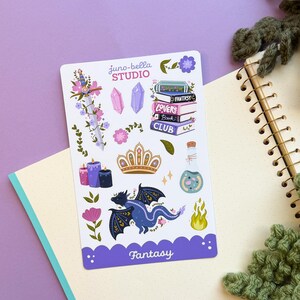 Puede incluir: Una hoja de pegatinas con ilustraciones de una espada con flores, cristales, una pila de libros con el texto Fantasy Lovers Book Club, una corona, velas, una poción, un dragón azul y una llama. El texto dice juno-bella STUDIO y Fantasy.