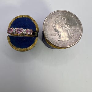 Puede incluir: Un anillo de oro con tres piedras preciosas rosas de corte ovalado y dos peque&ntilde;as piedras transparentes, exhibido en un soporte de terciopelo azul junto a una moneda de 25 centavos de d&oacute;lar para mostrar la escala de la joya.